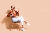 Full length photo of cheerful dreamy lady dressed brown top sitting chair showing looking empty space isolated beige color background