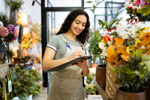 A young Latina girl with long hair works inside a flower shop dressed in an apron.The American woman is taking inventory with a notebook.Concept of working woman. Women's Day.