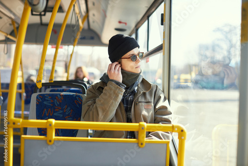 Young man, passenger in stylish clothes and sunglasses sitting in modern tram and listening to music in headphones. Concept of public transport, urban lifestyle