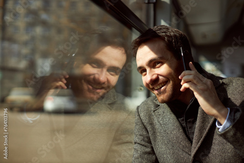 Close-up image of smiling man in grey pat using public transport, sitting in tram, talking on mobile phone and looking on window with smile. Sunny day. Concept of public transport, urban lifestyle
