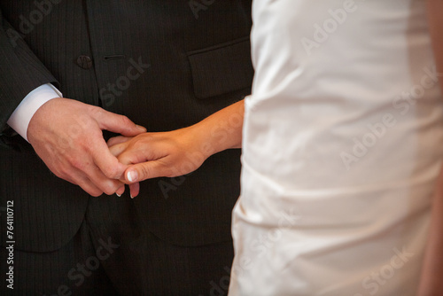 This close-up captures the tender moment where a bride places a wedding ring on the groom's finger. Their hands meet against the contrasting fabrics of the groom's suit and the bride's dress, a