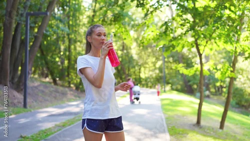 Tired young woman in sportswear drinking water from bottle and wiping sweat after jogging. Fit female athlete smiling and standing on footpath in summer park represents healthy lifestyle.