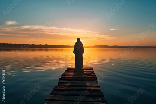 Silhouette of Jesus Christ at the edge of a dock, staring out at the sunrise over a calm lake.