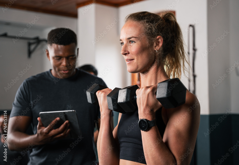 Woman lifts dumbbells in gym as man watches tablet - obrazy, fototapety, plakaty 