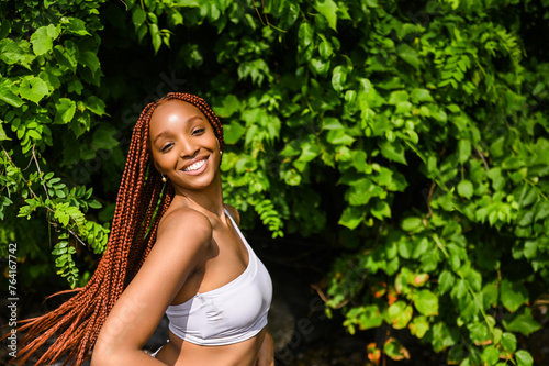 Outdoor portrait of natural Beautiful young African American woman long red braids hair style and perfect white teeth smile, posing in swimsuit at sunny summer day with green foliage beach background