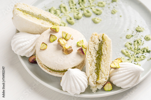 Close view of sweet crunchy macaron or french macaroon meringue-based confection with pistachio flavour ganache decorated with crushed nuts and cream cheese served on plate on white wooden table