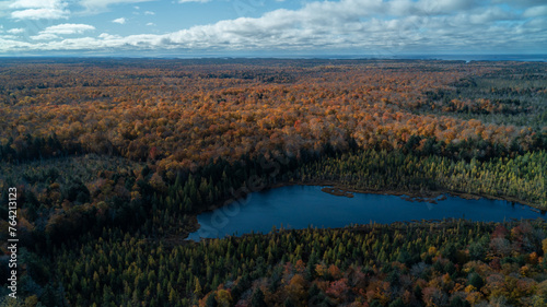 An aerial shot of a blue pond surrounded by a colorful forest in the fall on a sunny day