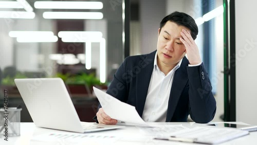 Overworked asian businessman is tired doing paperwork sitting at workplace in business office. A mature man in formal suit is overtired and bored. Overload exhausted financier has difficulties at work photo