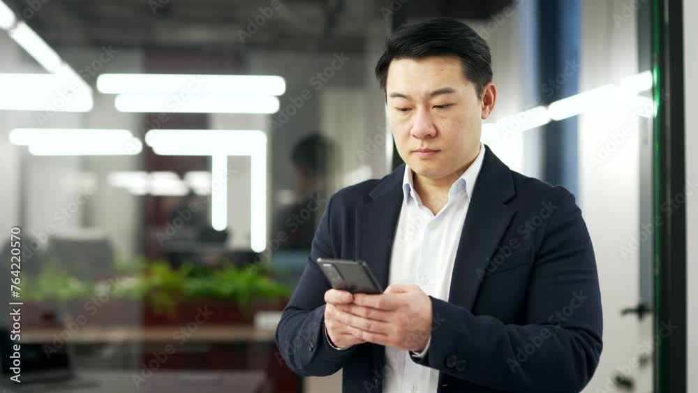 Serious asian businessman is using the phone while standing in business office. Focused man in formal suit reads or typing message, chats with client, banking or swiping browsing online on smartphone Serious asian businessman is using the phone while standing in business office. Focused man in formal suit reads or typing message, chats with client, banking or swiping browsing online on smartphone