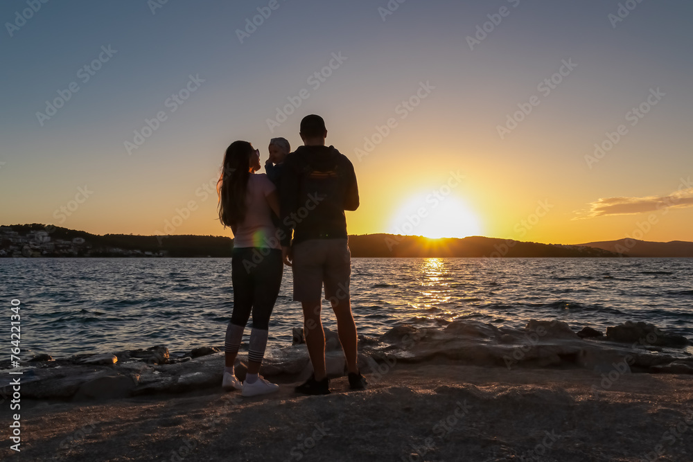 Silhouette of loving parents holding small toddler at romantic sunset in Okrug Gornij, Split-Dalmatia, Croatia. Coastline of island Otok Ciovo, Adriatic Sea in summer. Family travel vacation concept