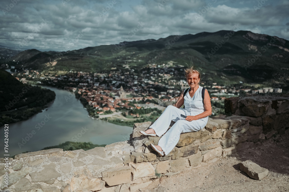 Fototapeta premium Mature woman tourist takes pictures on observation deck confluence of two rivers Aragvi and Kura, ancient city Mtskheta, Georgia. Retired people summer holiday vacation, active lifestyle concept. 