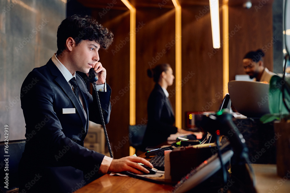 Hotel manager using computer while talking on the phone at reception desk.
