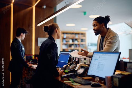 Quadro su tela Black man talking to receptionist while signing paperwork at hotel reception desk