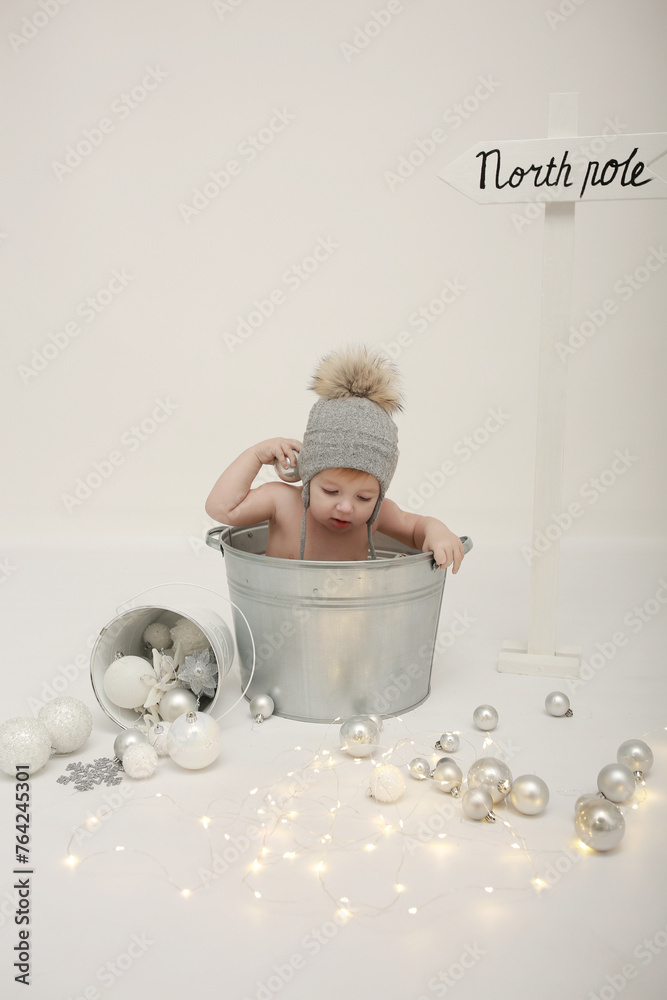 Naklejka premium a little boy sits in a metal basin on a white background in a cap. New Year's idea for photos of small children. scattered Christmas decorations. stylish children's photo zone. Merry Christmas 
