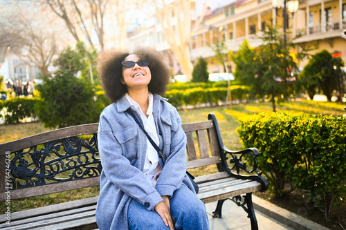 Fashion stylish portrait of attractive young natural beauty African American woman with afro hair in blue coat posing in nature park in green foliage. Perfect teeth smile. Happy lady in sun glasses