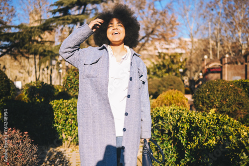 Close up Fashion street style portrait of attractive young natural beauty African American woman with afro hair in blue coat posing walking outdoors in sunny day. Happy lady with perfect teeth smile