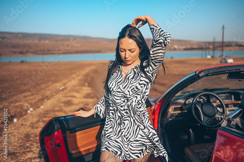 Happy smiling brunette woman driver in fashion dress posing with new red cabriolet car on beach coast, enjoying and ready for journey. Driving courses and life insurance concept. Glamorous lifestyle