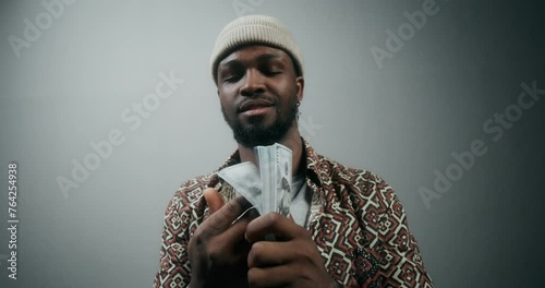 A stylish bearded man in a hat tosses a stack of money and smiles at the camera standing on a gray background in the studio photo