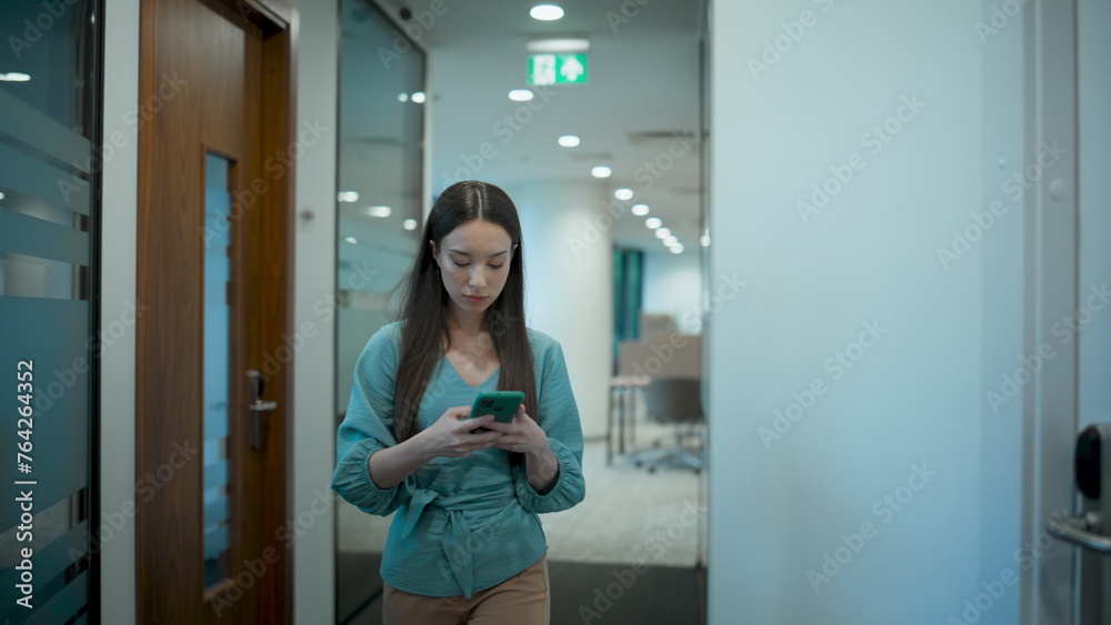 Asian businesswoman messaging mobile on go in office. Manager walking corridor