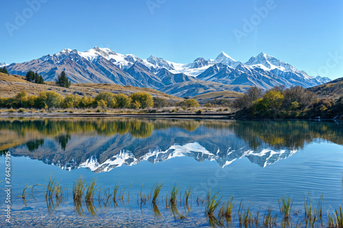A clear blue sky, snow-capped mountains, a calm lake, and some grass in the foreground..