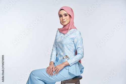 Beautiful female model wearing blue peplum dress with hijab, a modern lifestyle outfit for Muslim woman, sitting on a chair isolated over white background. Eidul fitri fashion and beauty concept. photo