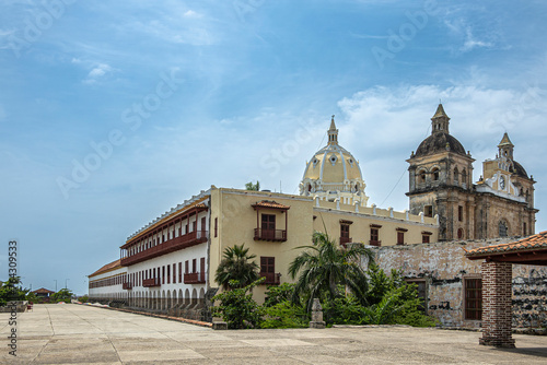 Cartagena, Colombia - July 25, 2023: Santuario de San Pedro Claver church towers, historic front facade with cloister and museum buildings on side under blue cloudscape. Green foliage in front photo