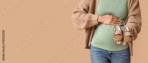 Young pregnant woman holding baby booties on brown background with space for text, closeup