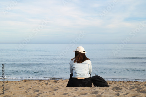 Girl with cap sitting on the seashore of a beach without people, feeling of serenity and solitude.
