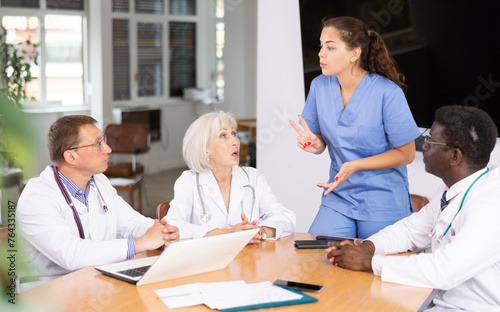 Group of different people medics in medical uniforms having discussion about work at table in office