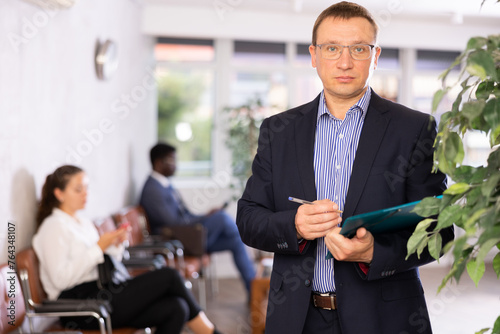 Dispassionate secretary man in glasses stands at entrance to office reception with folder and list of guests and expects invited visitors. Male office hostess willing to help