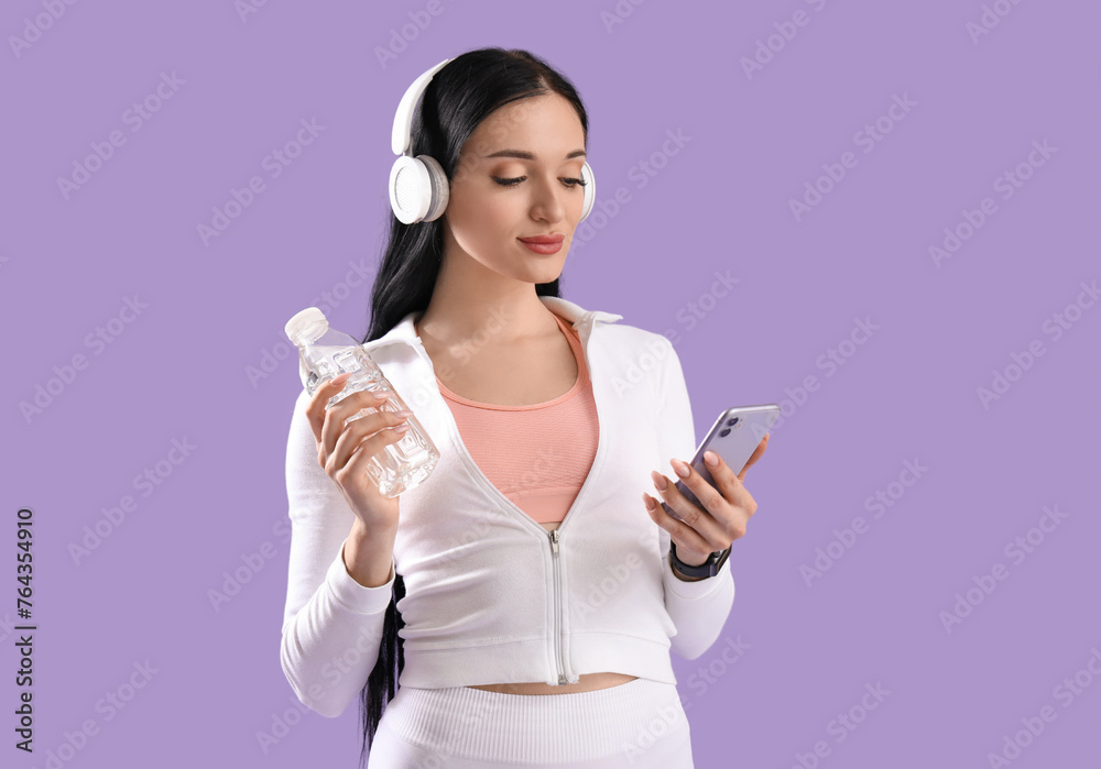 Portrait of sporty young woman with bottle of water, headphones and mobile phone on lilac background Portrait of sporty young woman with bottle of water, headphones and mobile phone on lilac background