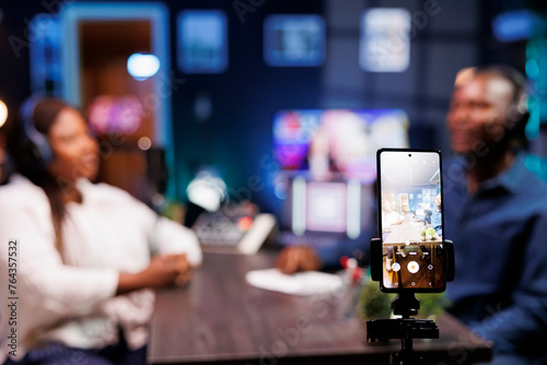 A vlogger is recording a podcast episode with a smartphone mounted on a tripod in a neon-lit studio. The show host is interviewing a guest and filming the discussion using a mobile device.