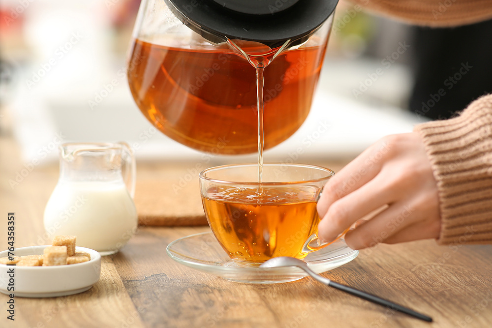 Young woman pouring black tea from teapot into cup in kitchen Young woman pouring black tea from teapot into cup in kitchen