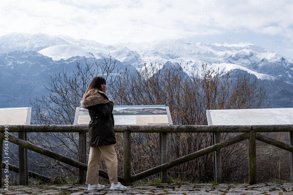 Young lady with brown hair, green coat, admires stunning snowy mountain vista.