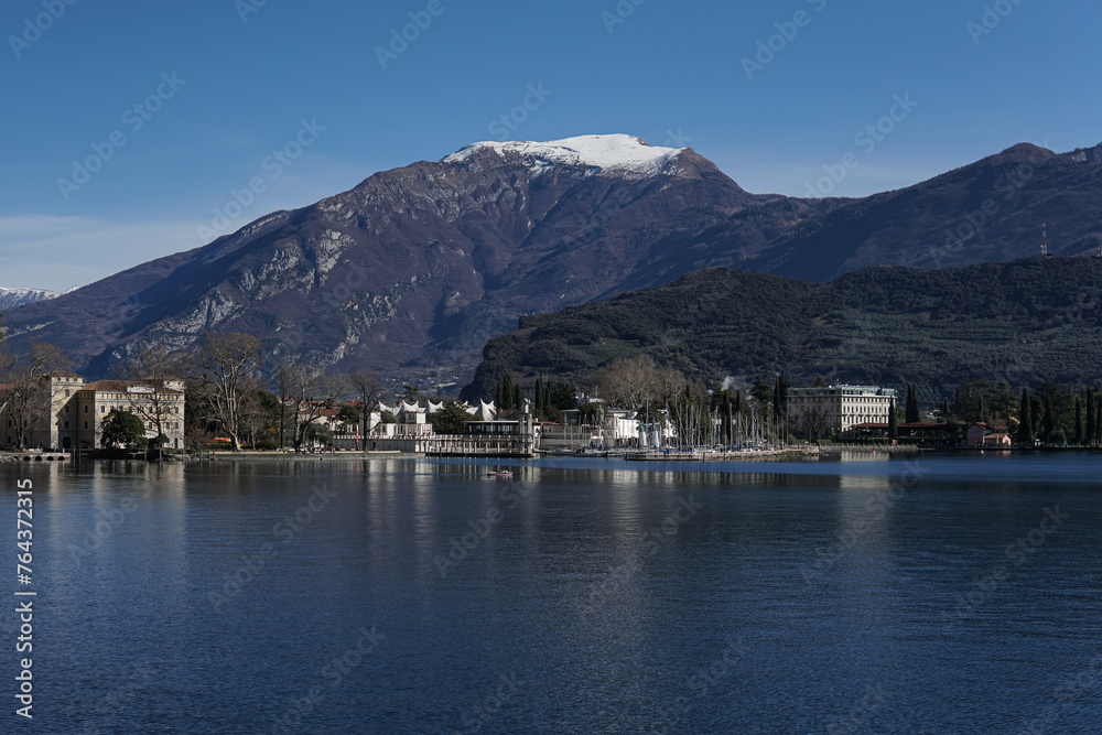 lake with the mountain in background, town on one side, buildings along shore line, clear blue sky, snow covered peak, small boats at dock, trees around water edge, calm waters