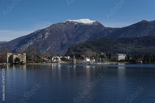 lake with the mountain in background, town on one side, buildings along shore line, clear blue sky, snow covered peak, small boats at dock, trees around water edge, calm waters