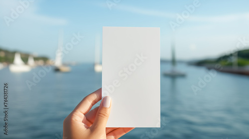 Female hand holding a piece of white paper on a blurred background of an ocean bay with yachts. Copy space for text.