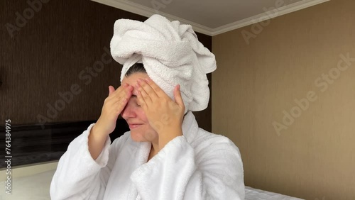 Horizontal 4K video. Young adult white woman in robe, towel on head applies moisturizer to face in hotel room. Skin protection during cold season. Cosmetic procedure in spa salon. Rest, relax, march 8