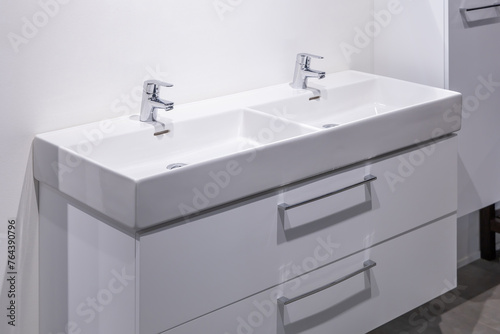Double white sink in a modern bathroom on a white cabinet with a smooth front and horizontal handles. Each sink has its own chrome faucet. Sleek design, minimalist aesthetics