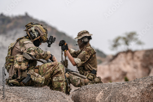 Soldiers in camo gear during a tactical operation with binoculars and rifles on a rocky terrain, showcasing military precision and readiness.