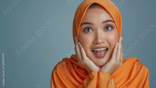 Excited woman in orange hijab smiling - A joyful woman with her hands on her cheeks wearing an orange hijab poses with an excited expression