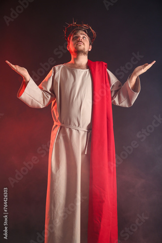 Man in Jesus robe and crown of thorns outstretching hands with smoke on black background