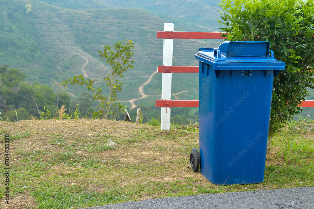 Beside the hill, on the roadside, sits a plastic blue garbage container. Beside the hill, on the roadside, sits a plastic blue garbage container.