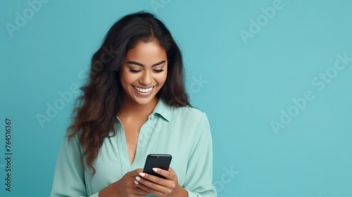 Young adult smiling happy pretty latin woman holding mobile phone looking at smartphone, typing message doing ecommerce shopping on cell, using trendy apps on cellphone isolated on blue background