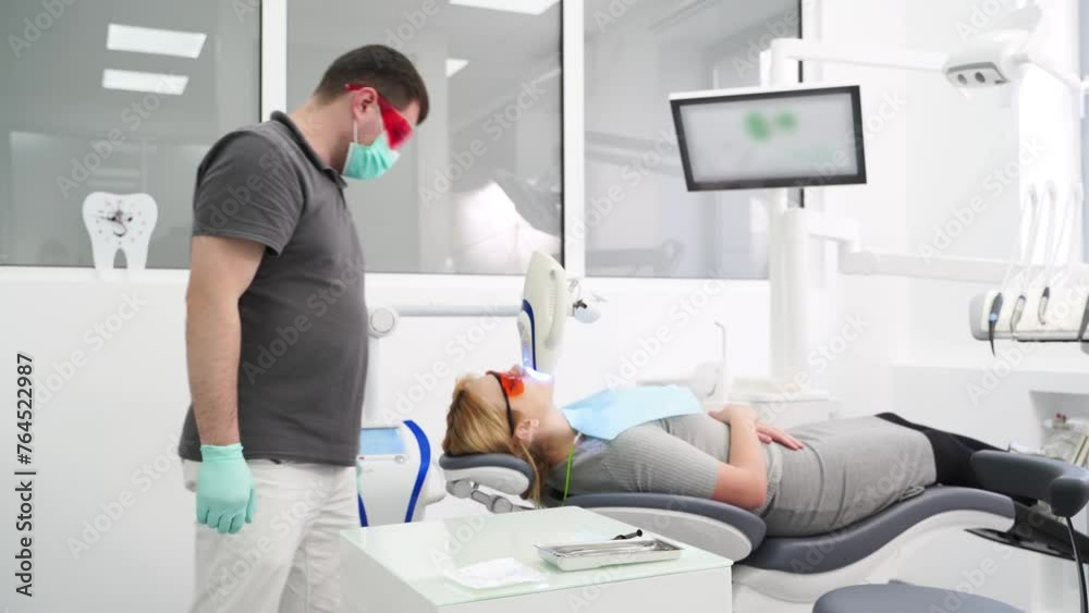 Professional UV whitening teeth procedure in the modern dental clinic. Male dentist checking female patient during laser teeth bleaching. Blonde woman in protective red glasses. Cosmetic dentistry. Professional UV whitening teeth procedure in the modern dental clinic. Male dentist checking female patient during laser teeth bleaching. Blonde woman in protective red glasses. Cosmetic dentistry.