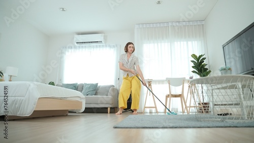 Caucasian housekeeper mopping the floor while dancing in lively mood at modern apartment. Happy energetic mother or cleaner enjoy cleaning the living room while moving to music. Lifestyle. Pedagogy.