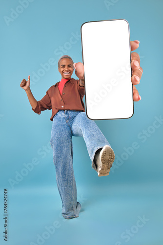 Young energetic excited African American woman laughing showing new smartphone with blank white screen and demonstrates thumbs up gesture making leg swing stands in turquoise studio. Mock up, template
