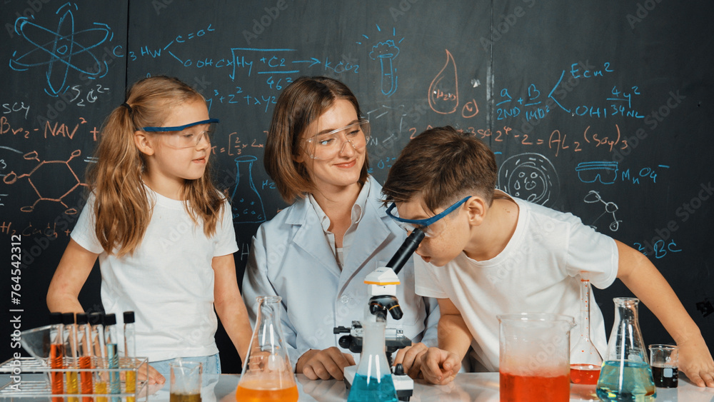 Caucasian boy looking under microscope while teacher giving advice. Professional instructor wearing lab suit looking for diverse student at table with beaker filled with colored solution. Erudition. - obrazy, fototapety, plakaty 