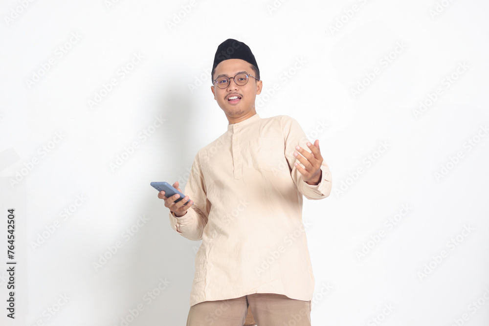 Portrait of surprised Asian muslim man in koko shirt with skullcap holding mobile phone, showing wow shocked face expression. Advertising and social media concept. Isolated image on white background