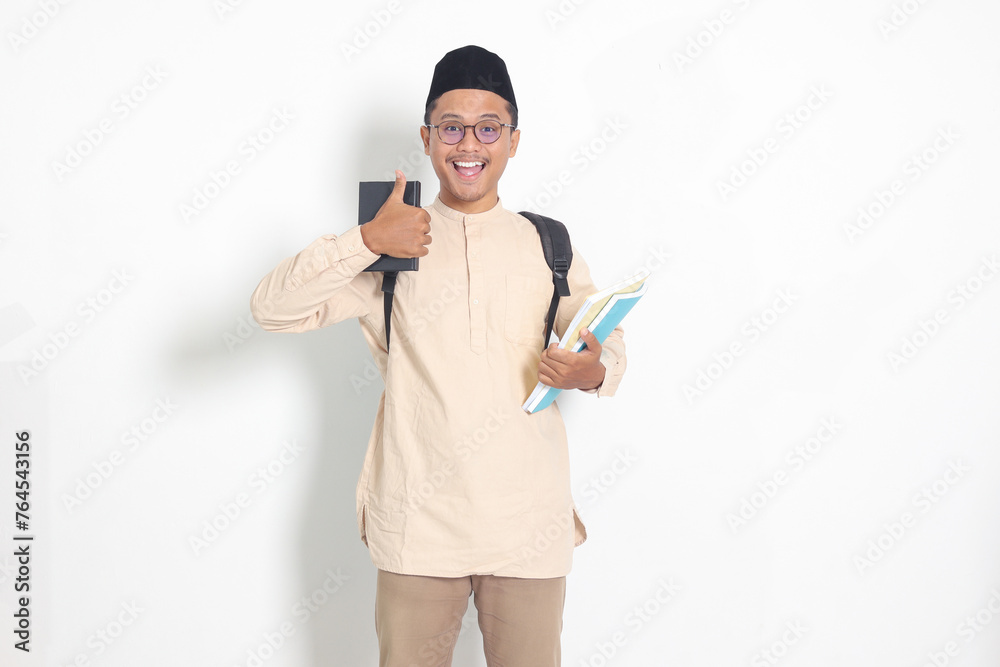 Portrait of excited student Asian muslim man in koko shirt with skullcap carrying backpack, holding school book, showing thumb up gesture. Islamic education concept. Isolated image on white background Portrait of excited student Asian muslim man in koko shirt with skullcap carrying backpack, holding school book, showing thumb up gesture. Islamic education concept. Isolated image on white background